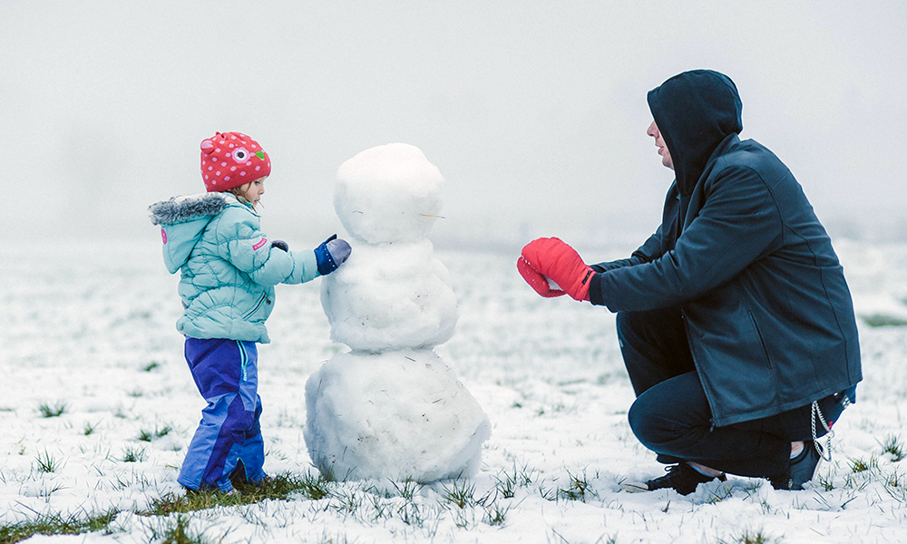 father and child making a snowman in snow-covered grassy area