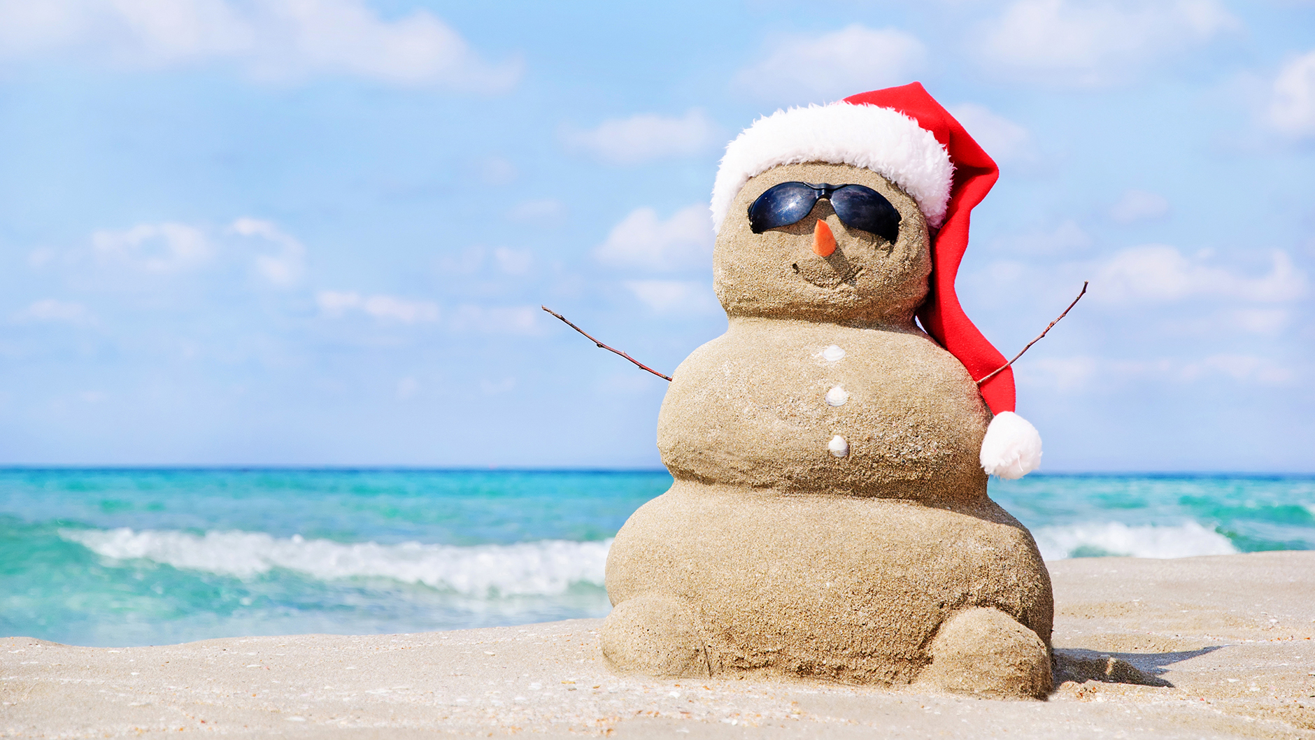 sand snowman on beach waring sunglasses and Santa hat with surf in background