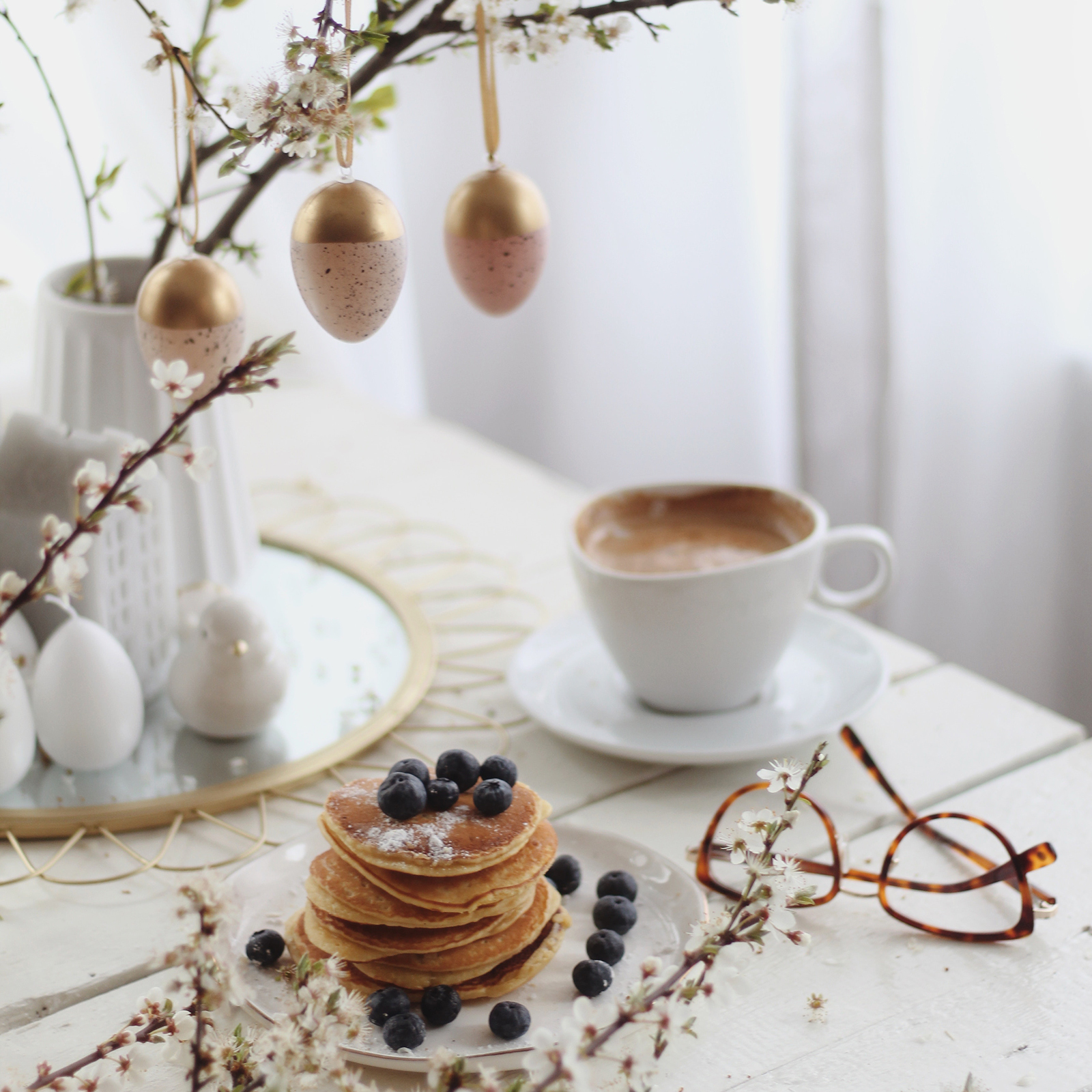 table set for Easter brunch with hot drink, pancakes, branches with gold egg decorations