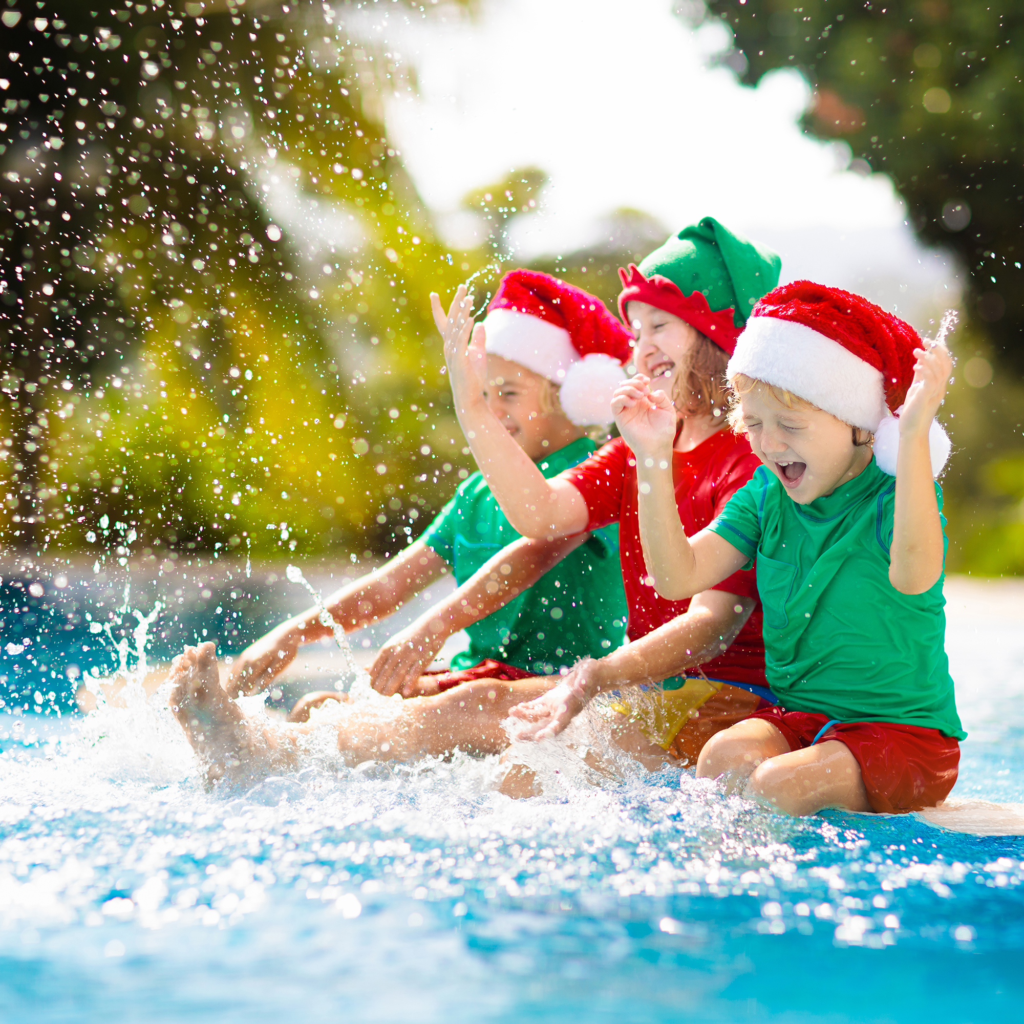 three kids dressed in green and red, wearing Santa and elf hats, kicking water from the edge of a swimming pool