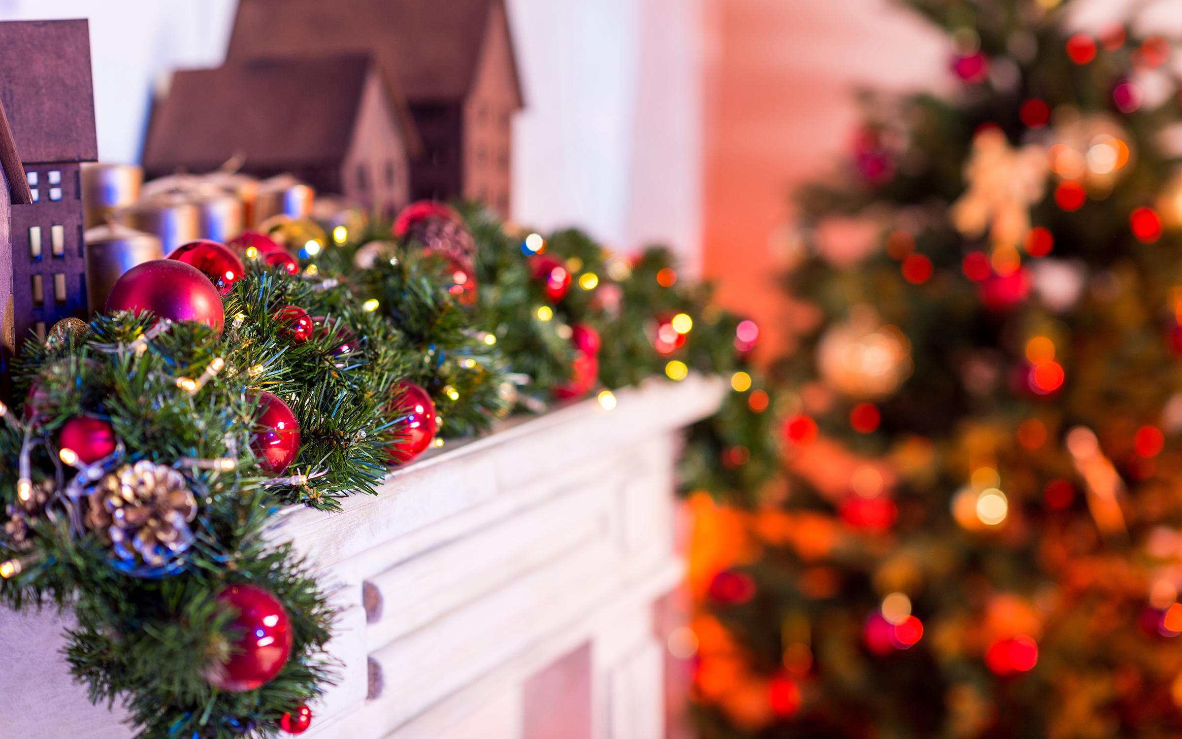 mantel decorated with pine garland, ornaments, wooden Christmas village buildings, Christmas tree in background