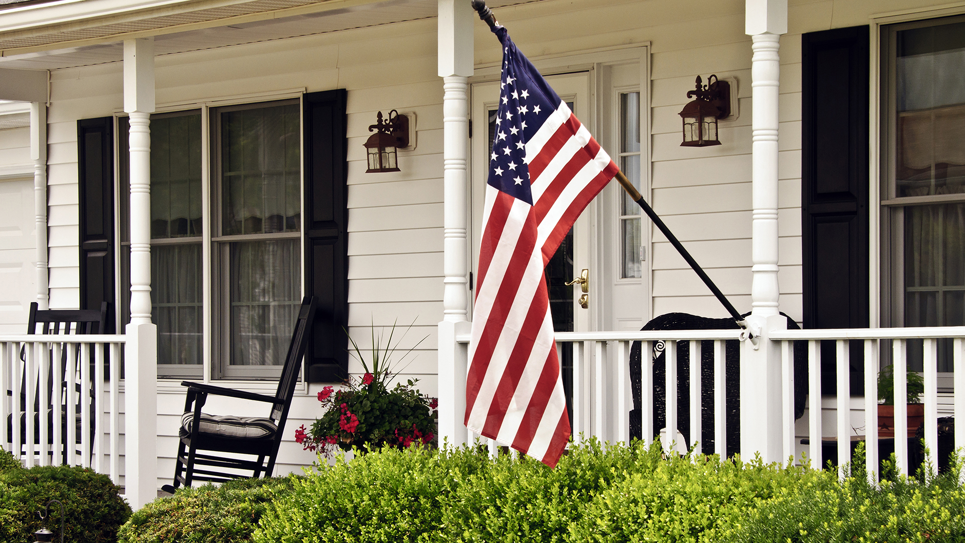 American flag hanging in front of white house with wide front porch decorated with flowers and rocking chairs